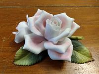 Ceramic pink rose with delicate petals and green leaves on a wooden surface.