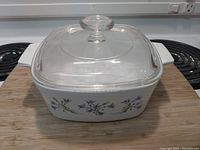 Square white ceramic Corning Ware baking dish with blue floral pattern and clear glass lid on stove.