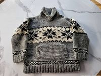 Full view of the grey Cowichan style sweater showing the shawl collar, kangaroo pocket, and black and white geometric pattern band across chest and sleeves.