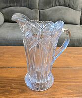 Photo showing front view of a clear glass pitcher with intricate cut glass pattern on a wooden surface, light reflecting on the glass.