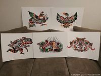 Photo showing five Indigenous style art prints by John Nelson placed on a sofa: Thunderbird, Eagle, Sea Monster, Otter, Sea Urchin, and Longhouse Mask designs in vibrant color palette.