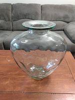 Photo of a large clear glass vase placed on a wooden table with a gray couch in the background. The vase has a rounded bulbous body and a wide flared rim, without any chips or cracks visible.