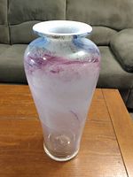 Photo of a tall glass vase on a wooden table with a brown couch in the background. The vase is mostly translucent with purple swirled patterns and a flared top rim.