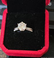 Front-facing image of the sterling silver ring with a large central Moissanite stone and accent stones on the band set inside a red velvet-lined black box.