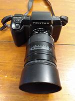 Front view of Pentax SF10 camera with Sigma zoom lens and lens hood attached on wooden surface.