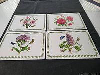 All 4 floral design Pimpernel place mats laid out on a black cloth, showing detailed botanical flower illustrations with butterflies and green leaf border.