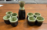 Ceramic saki set including one carafe and eight cups, set on a wooden surface with a grey sofa background.