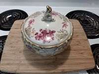 Closed view of the porcelain serving bowl with lid, decorated with pink floral and gold accents, placed on a wooden board.