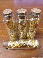Four glass vials containing gold leaf flakes sealed with cork stoppers, arranged standing and one laid horizontally.