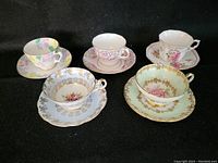 Five assorted vintage fine china teacups and saucers arranged on black background, showing various floral patterns and gold trims.