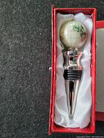 Wine stopper with ceramic globe design and metal body placed inside a red padded box with white satin lining.