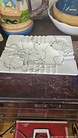White rectangular porcelain ashtray with abstract black line design on wooden surface alongside other ashtrays and items.
