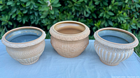 Photo of three different light-toned Roman style clay plant pots on a white surface outdoors with greenery background. Each is approximately 10.5" diameter and 8" height with glazed interiors and unique molded designs.