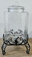 Front view of clear hammered glass drink dispenser on decorative black metal stand. Shows lid with round handle and pour spout at base.