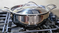 Side view of shiny stainless steel covered casserole on gas stove showing polished finish and clear glass lid.