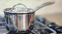 Side view of the stainless steel covered saucepan placed on a stove burner, showing the handle, lid, and polished reflective surface.