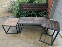 Wood and metal yard porch bench with back and two accompanying wood and metal tables on concrete floor outside.