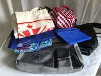 Photo showing the collection of aprons and various handbags including a white canvas tote with Canada design, a blue apron, a red and white checkered cosmetic bag, black tote bags, and a black handbag with floral embossing.