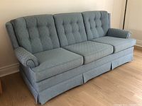 Angled front-left view of the light blue sofa showing tufted back cushions, rolled armrests, and skirted base.
