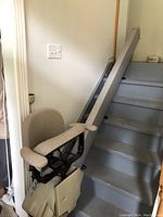 Photo showing Savaria stair lift chair at the base of indoor stairs with beige fabric seat and armrests, metal rail system installed along seven-step staircase.