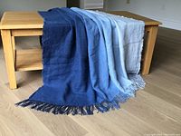 Ombre linen throw draped on wooden coffee table showing full gradient from deep indigo to light sky blue with fringed edges.