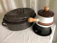 Black and white speckled oval roaster with lid and vintage off-white fondue pot with warming base shown together on a white surface.