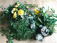 Top-down view of assorted artificial flowers and greenery in a box, showing yellow roses and multiple types of foliage.