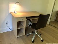 Front angled view of wood-effect desk with pull-out keyboard tray, open shelving, brown bentwood office chair on caster wheels, and silver-tone halogen desk lamp turned on