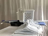 Photo of entire lot showing Rubbermaid small laundry hamper, ironing board with blue cover, white IKEA plastic bag holder, white and silver curtain rods, and plastic clothes hangers stacked in front.