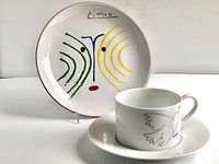 Porcelain cup and matching saucer with Picasso dove design and signature, placed in front of stoneware side plate with colorful abstract face artwork and red rim.