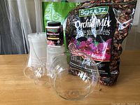 Photo showing clear plastic orchid pots stacked, bottle of Schultz Orchid Food, partial bag of Schultz Orchid Mix growing medium, and three clear glass hanging holders for air plants of different sizes