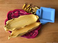 Photo showing a stack of 6 burgundy crab-shaped plates, 6 creamy yellow corn cob-shaped plates, and 6 light blue square small plates along with 14 yellow corn cob holders in two designs.