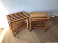 Photo of two solid wood end tables side by side against a white wall on wooden floor, showing slatted tops and lower shelves.