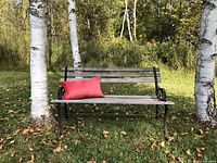 Outdoor rustic bench set on grass with birch trees in background, showing full bench and coral red pillow on left side