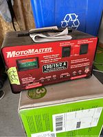 Rectangular black and red MotoMaster battery charger with voltage and function selectors, and attached clamps and cables.
