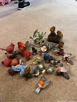 Photo showing an assortment of bird figurines in various colors and species arranged on carpeted floor, including cardinals, blue jays, robins, and others, some hanging and some standing, plus a green succulent decor piece.
