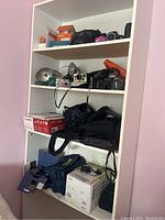 White four-shelf bookcase with assorted cameras, flashes, film boxes, bags, printer box and cables