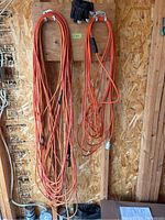 Two orange extension cords hanging side-by-side on a plywood wall with a pair of gloves above them.