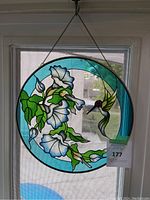 Hanging circular stained glass panel with blue border, white flowers, green leaves and a hummingbird, hung by chain on window.