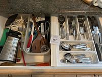 View into two open kitchen drawers showing a variety of kitchen utensils including cutlery such as forks, spoons, and knives, as well as cooking tools like graters, peelers, whisks, spatulas, and scissors.