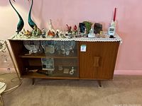 Side view of MCM style display cabinet with decorative items on top and inside, showing legs, sliding glass door, and wooden door with handle.