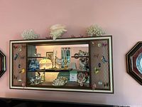 Front view of the MCM wall-mounted display shelf showing wood frame, metal mesh side panels, glass shelves, mirror back, and decorative items inside.