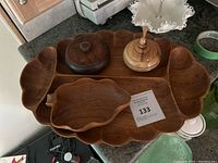 Photo showing a collection of wooden serving pieces on a countertop including a large scalloped tray, leaf-shaped bowl, round lidded container, and small turned wood container with lid and handle