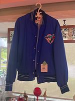Front view of the men's vintage Blue Jays bomber jacket hanging, showing royal blue color, ribbed collar, cuffs and waistband with stripes, front button closure, and Blue Jays logo patch on chest.