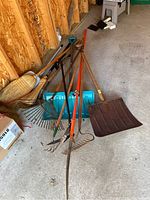 Full set of garden tools including leaf rake, snow shovel, pruning shears, metal rakes, broom, and plastic shovel shown grouped on concrete floor against wooden wall.