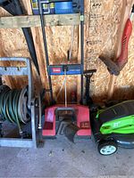 Red Toro Power Clear snowblower with black discharge chute and vertical handles, stored indoors beside garden hose and electric lawn mower.