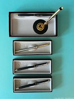 Five Quill salesman sample pens displayed in felt-lined black presentation boxes alongside a desk pen and base stand on a turquoise background.
