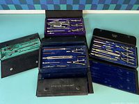 Three antique drafting kits displayed open showing leather cases with blue and purple velvet interiors and assorted drafting tools including compasses, dividers, and ruling pens.