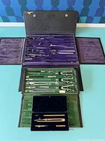 Open large black drafting tool case showing multiple compasses, dividers, ruling pens, beam compasses in velvet-lined compartments