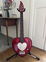 Front view of red heart-shaped electric guitar on stand showing full body and neck.